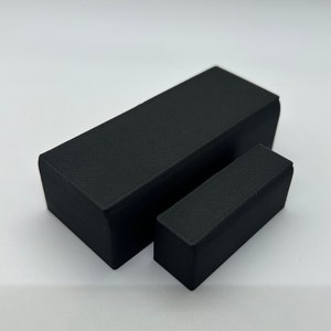 May include: Two black rectangular-shaped objects, one larger than the other, made of a textured material. The objects are on a white surface.