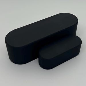 May include: Two matte black, rectangular, rounded-edge boxes of different sizes. The larger box is positioned behind the smaller one, both resting on a light gray surface. The boxes have a smooth, solid appearance.