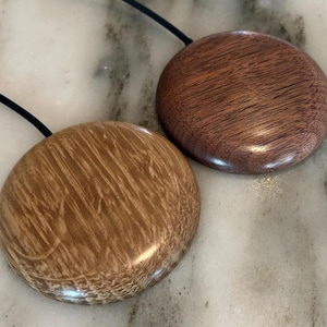 Pode incluir: Dois colares com pingentes de madeira. Um pingente é marrom claro com um padrão de grão único, e o outro é marrom mais escuro com um acabamento liso. Ambos são circulares e presos a cordões pretos. Os pingentes são exibidos em uma superfície de mármore.