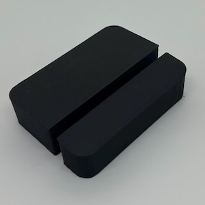 May include: A black phone stand with a rectangular shape and rounded corners. The stand is divided into two parts, with a slot in between to hold a phone or tablet. The stand is made of a solid, matte material.