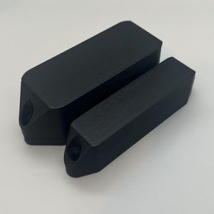 May include: Two black rectangular objects with a textured surface. The larger object is positioned behind the smaller one. Each object has a rounded end with a hole. The objects are on a white surface.