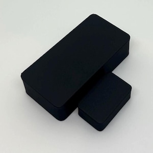 May include: Two black rectangular boxes of different sizes are displayed against a white background. The boxes have a matte finish and appear to be made of a solid material. The larger box is positioned diagonally, with the smaller box beside it.
