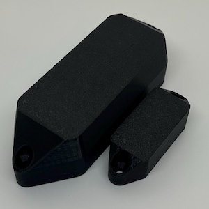 May include: Two black, textured, geometric objects. The larger one is rectangular with a pointed end and a hole. The smaller one has a similar design. Both have a matte finish and appear to be 3D-printed.