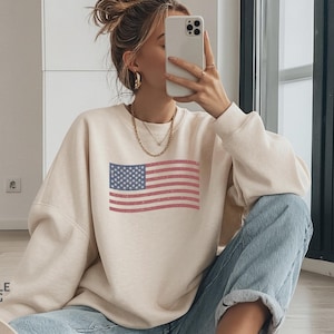 May include: A sand-colored crewneck sweatshirt featuring a distressed-style American flag graphic on the chest, paired with light blue denim jeans and white canvas sneakers. The sweatshirt has long sleeves and a relaxed fit.