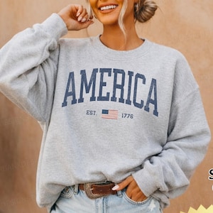 May include: A light grey crewneck sweatshirt featuring the word AMERICA in large dark blue capital letters, with the text EST. 1776 and a small American flag graphic centered below. The sweatshirt has long sleeves and a ribbed collar and cuffs.