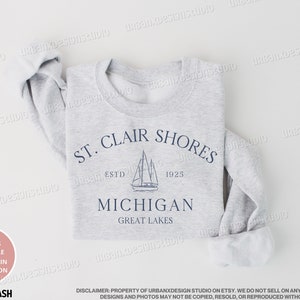 May include: A gray sweatshirt with a navy blue graphic design that says "St. Clair Shores, Est. 1925, Michigan, Great Lakes". The graphic also includes a sailboat.