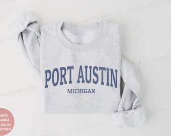 Puede incluir: Sudadera gris claro con el texto "PORT AUSTIN MICHIGAN" en azul marino. La sudadera está doblada cuidadosamente, con las mangas enrolladas. Diseño sencillo y clásico.
