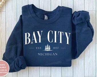 Puede incluir: Una sudadera azul marino con texto blanco que dice "Bay City - Est. 1837 - Michigan". El texto está rodeado por una línea blanca y un pequeño gráfico de pinos.