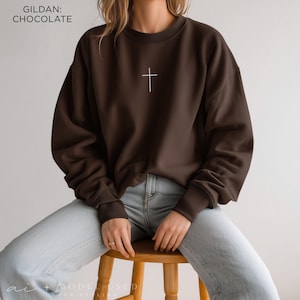 Puede incluir: Una sudadera de cuello redondo Gildan en color marr&oacute;n chocolate con un peque&ntilde;o gr&aacute;fico de una cruz blanca centrado en el pecho. El jersey de manga larga tiene cuello, pu&ntilde;os y dobladillo acanalados, combinado con vaqueros azul claro.