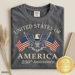 Puede incluir: Una camiseta gris carb&oacute;n con un gr&aacute;fico de un &aacute;guila calva centrado entre dos banderas estadounidenses. El texto UNITED STATES OF AMERICA y 250th Anniversary aparece en color crema. Incluye la etiqueta de cuello de la marca Comfort Colors.