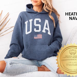 May include: A heather navy blue hooded sweatshirt featuring the text USA in large white block letters across the chest, with a small American flag graphic positioned directly below the text. The hoodie includes a front kangaroo pocket.