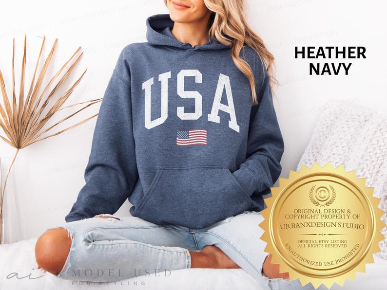 May include: A heather navy blue hooded sweatshirt featuring the text USA in large white block letters across the chest, with a small American flag graphic positioned directly below the text. The hoodie includes a front kangaroo pocket.