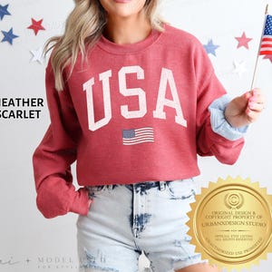 May include: A heather scarlet crewneck sweatshirt featuring the text USA in large white block letters with a distressed finish. Below the text is a small graphic of the American flag. The sweatshirt has long sleeves and a ribbed neckline.