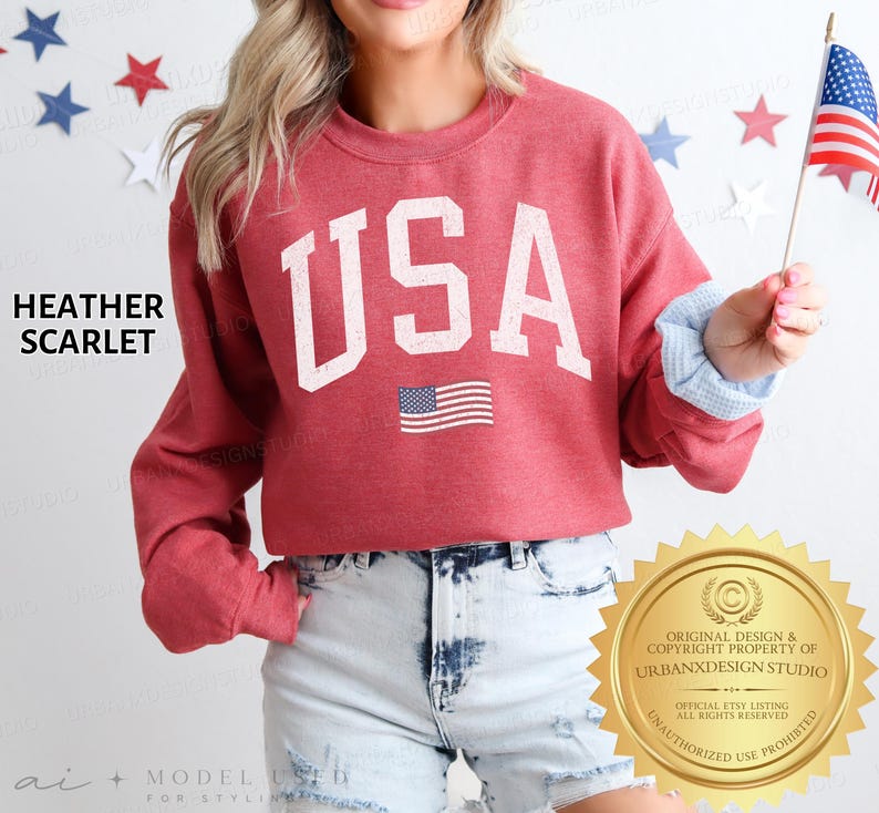 May include: A heather scarlet crewneck sweatshirt featuring the text USA in large white block letters with a distressed finish. Below the text is a small graphic of the American flag. The sweatshirt has long sleeves and a ribbed neckline.
