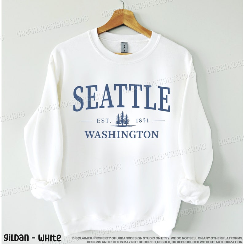 May include: White crewneck sweatshirt with a blue graphic design that says "Seattle Est. 1851 Washington" with a silhouette of pine trees.