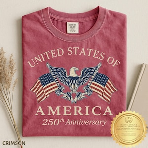Puede incluir: Una camiseta de cuello redondo color rojo carmes&iacute; con un gr&aacute;fico de un &aacute;guila calva con las alas extendidas, flanqueada por dos banderas estadounidenses. El texto dice UNITED STATES OF AMERICA 250th Anniversary. Etiqueta Comfort Colors en el cuello.