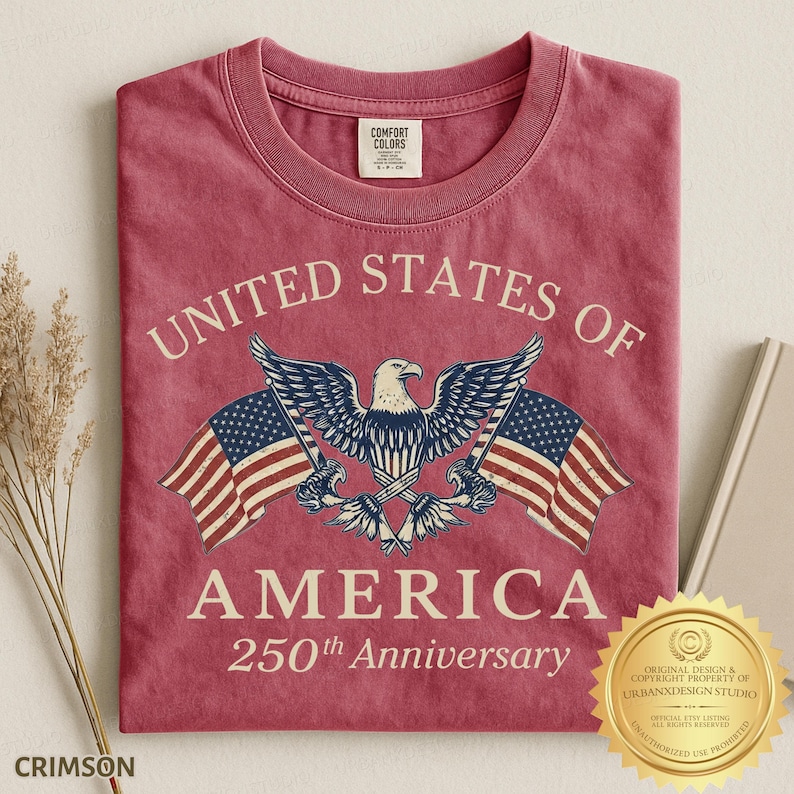 Puede incluir: Una camiseta de cuello redondo color rojo carmes&iacute; con un gr&aacute;fico de un &aacute;guila calva con las alas extendidas, flanqueada por dos banderas estadounidenses. El texto dice UNITED STATES OF AMERICA 250th Anniversary. Etiqueta Comfort Colors en el cuello.