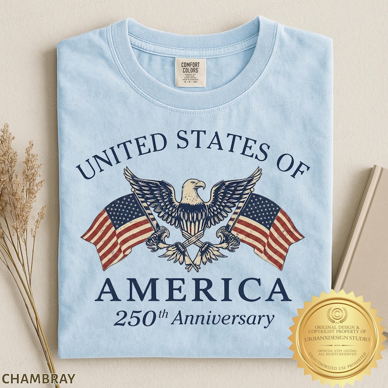 Puede incluir: Una camiseta azul claro con un gr&aacute;fico de un &aacute;guila calva centrado entre dos banderas estadounidenses. El texto dice UNITED STATES OF AMERICA y 250th Anniversary en letras azul oscuro. La camiseta tiene una etiqueta de cuello Comfort Colors.