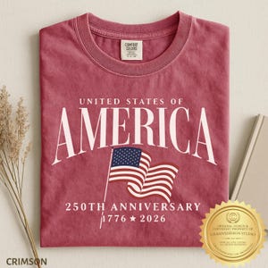 May include: A crimson red crew neck t-shirt featuring the white text UNITED STATES OF AMERICA, a graphic of a waving American flag, and the text 250TH ANNIVERSARY 1776 2026. The shirt has a Comfort Colors brand label on the inside of the collar.