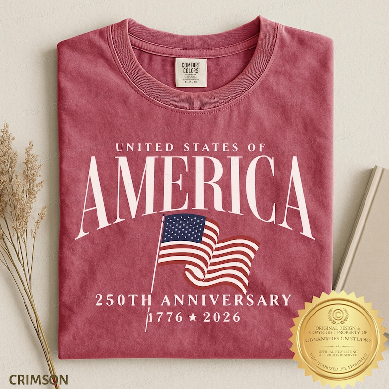 May include: A crimson red crew neck t-shirt featuring the white text UNITED STATES OF AMERICA, a graphic of a waving American flag, and the text 250TH ANNIVERSARY 1776 2026. The shirt has a Comfort Colors brand label on the inside of the collar.