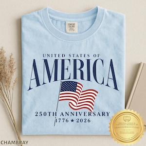 May include: A light blue t-shirt featuring the dark blue text UNITED STATES OF AMERICA, a graphic of the American flag, and the text 250TH ANNIVERSARY 1776 2026. The shirt has a Comfort Colors brand label on the inside of the crew neckline.