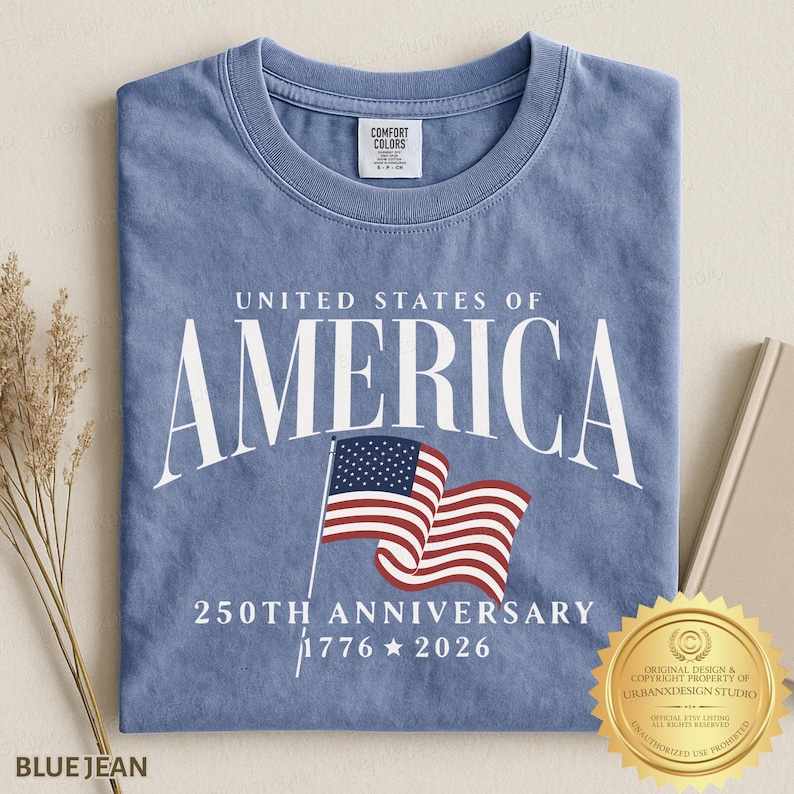 May include: A blue crew neck t-shirt featuring the white text UNITED STATES OF AMERICA, a graphic of a waving American flag, and the text 250TH ANNIVERSARY 1776 2026. The shirt has a Comfort Colors brand label on the inside of the collar.