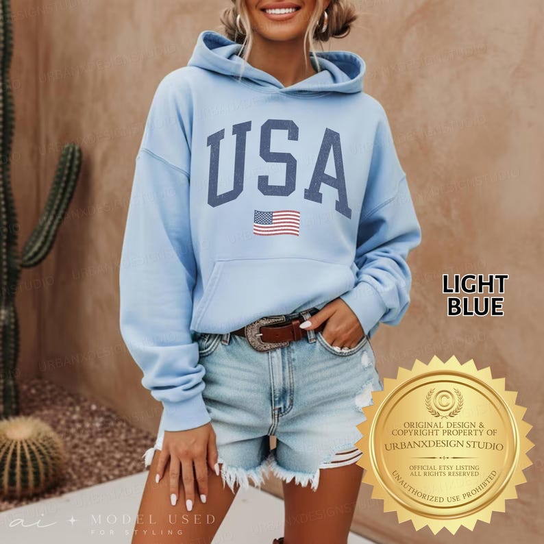 May include: A light blue hooded sweatshirt featuring the text USA in large dark blue block letters across the chest, accompanied by a small American flag graphic positioned directly below the text. The garment includes a front kangaroo pocket.