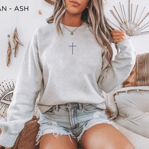 Puede incluir: Una sudadera de cuello redondo Gildan en color gris ceniza con un peque&ntilde;o gr&aacute;fico de una cruz negra centrado en el pecho. El jersey de manga larga tiene cuello, pu&ntilde;os y dobladillo acanalados, y se combina con pantalones cortos de mezclilla.
