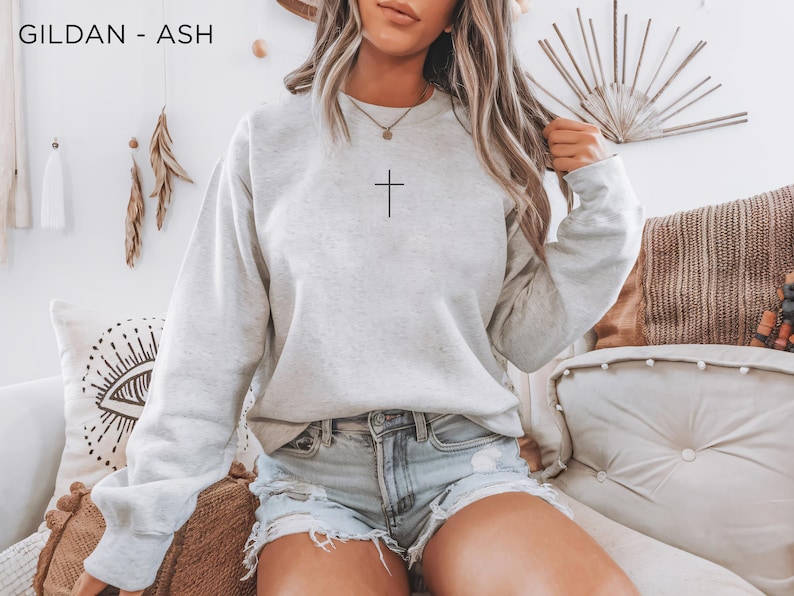 Puede incluir: Una sudadera de cuello redondo Gildan en color gris ceniza con un peque&ntilde;o gr&aacute;fico de una cruz negra centrado en el pecho. El jersey de manga larga tiene cuello, pu&ntilde;os y dobladillo acanalados, y se combina con pantalones cortos de mezclilla.