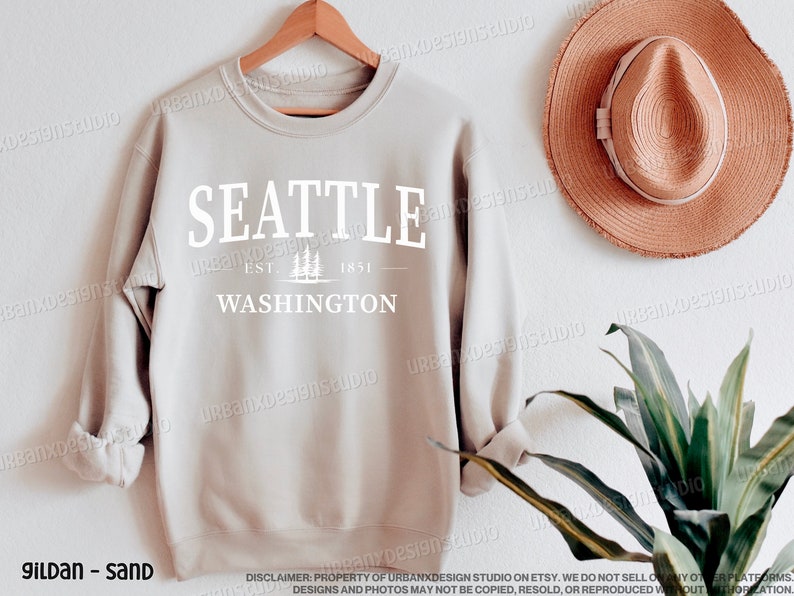 May include: A sand-colored sweatshirt with the text "SEATTLE EST. 1851 WASHINGTON" printed in white on the front. The sweatshirt is hanging on a wooden hanger against a white wall.