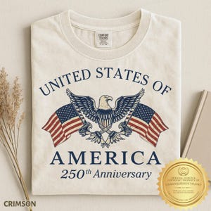 Puede incluir: Una camiseta de color crema con un gr&aacute;fico de un &aacute;guila calva centrada entre dos banderas estadounidenses. El texto UNITED STATES OF AMERICA 250th Anniversary aparece en letras azul oscuro. La camiseta tiene una etiqueta de cuello Comfort Colors.