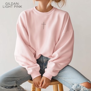 Puede incluir: Una sudadera de cuello redondo Gildan en color rosa claro con un peque&ntilde;o gr&aacute;fico de una cruz negra centrado en el pecho. El jersey de manga larga tiene cuello, pu&ntilde;os y dobladillo acanalados, combinado con vaqueros de mezclilla azul claro.