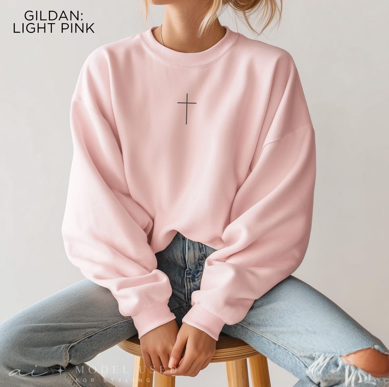 Puede incluir: Una sudadera de cuello redondo Gildan en color rosa claro con un peque&ntilde;o gr&aacute;fico de una cruz negra centrado en el pecho. El jersey de manga larga tiene cuello, pu&ntilde;os y dobladillo acanalados, combinado con vaqueros de mezclilla azul claro.