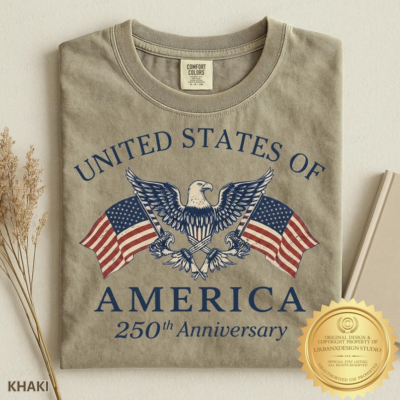 Puede incluir: Una camiseta de cuello redondo color caqui con un gr&aacute;fico de un &aacute;guila calva entre dos banderas estadounidenses. El texto UNITED STATES OF AMERICA 250th Anniversary aparece en letras azul oscuro. Etiqueta de cuello Comfort Colors visible.