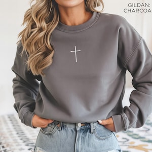 Puede incluir: Una sudadera de cuello redondo en color gris carb&oacute;n con un peque&ntilde;o y sencillo gr&aacute;fico de una cruz blanca centrado en el pecho. La prenda tiene mangas largas con pu&ntilde;os acanalados y cuello acanalado. El texto GILDAN: CHARCOAL aparece arriba a la derecha.