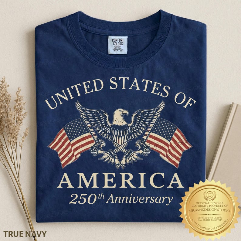 Puede incluir: Una camiseta azul marino con un gr&aacute;fico de un &aacute;guila calva centrado entre dos banderas estadounidenses. El texto UNITED STATES OF AMERICA 250th Anniversary aparece en color crema. La camiseta tiene una etiqueta de la marca Comfort Colors.