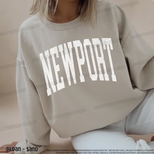 May include: A light beige sweatshirt with the word "NEWPORT" printed in white on the front. The sweatshirt is made of a soft, comfortable material and has a relaxed fit.