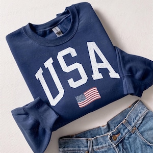 May include: A navy blue crewneck sweatshirt featuring the white distressed text USA and a small American flag graphic on the front. The sweatshirt is paired with blue denim jeans that have a small rectangular red and white label on the pocket.