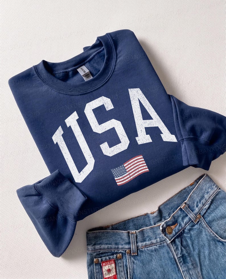May include: A navy blue crewneck sweatshirt featuring the white distressed text USA and a small American flag graphic on the front. The sweatshirt is paired with blue denim jeans that have a small rectangular red and white label on the pocket.