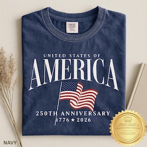 May include: A navy blue crew neck t-shirt featuring the white text UNITED STATES OF AMERICA, a graphic of a waving American flag, and the text 250TH ANNIVERSARY 1776 2026. The shirt has a Comfort Colors brand label on the inside of the collar.