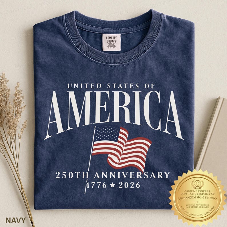 May include: A navy blue crew neck t-shirt featuring the white text UNITED STATES OF AMERICA, a graphic of a waving American flag, and the text 250TH ANNIVERSARY 1776 2026. The shirt has a Comfort Colors brand label on the inside of the collar.
