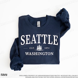 May include: A navy blue sweatshirt with white text that reads "SEATTLE - EST. 1851 - WASHINGTON" with a graphic of pine trees.