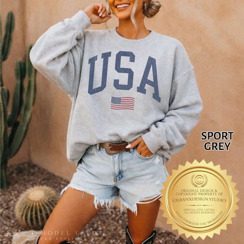 May include: A sport grey crewneck sweatshirt featuring the text USA in large navy blue block letters with a small American flag graphic centred below the text. The sweatshirt has long sleeves and a ribbed neckline, cuffs, and hem.
