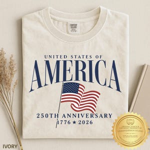 May include: An ivory crew neck t-shirt featuring the dark blue text UNITED STATES OF AMERICA, a graphic of the American flag, and the text 250TH ANNIVERSARY 1776 2026. The shirt has a Comfort Colors brand label on the inside of the collar.
