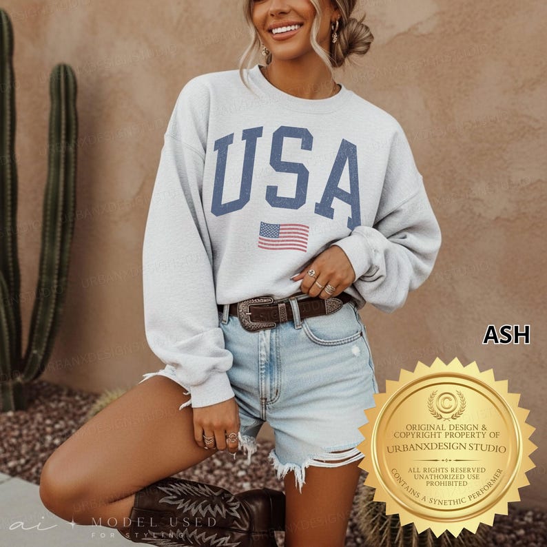 May include: A light grey crewneck sweatshirt featuring the text USA in large blue block letters with a small American flag graphic centred below. The sweatshirt has long sleeves and a relaxed fit, paired with light blue denim shorts and a brown belt.