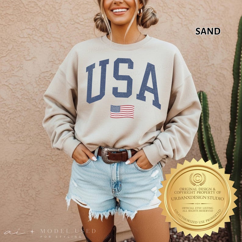May include: A sand-coloured crewneck sweatshirt featuring the text USA in large, dark blue block letters with a distressed texture. Below the text is a small, rectangular American flag graphic. The sweatshirt has long sleeves and a ribbed neckline.