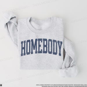 May include: A gray sweatshirt with the text "HOMEBODY" printed in navy blue letters. The sweatshirt is folded and laid flat on a white surface.