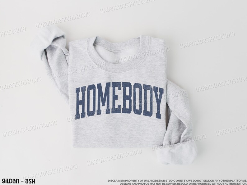May include: A gray sweatshirt with the text "HOMEBODY" printed in navy blue letters. The sweatshirt is folded and laid flat on a white surface.