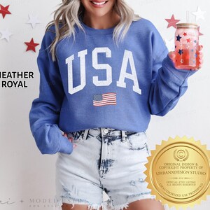 May include: A heather royal blue crewneck sweatshirt featuring the text USA in large white block letters with a distressed finish and a small American flag graphic below. The sweatshirt is paired with light blue acid-wash denim shorts.