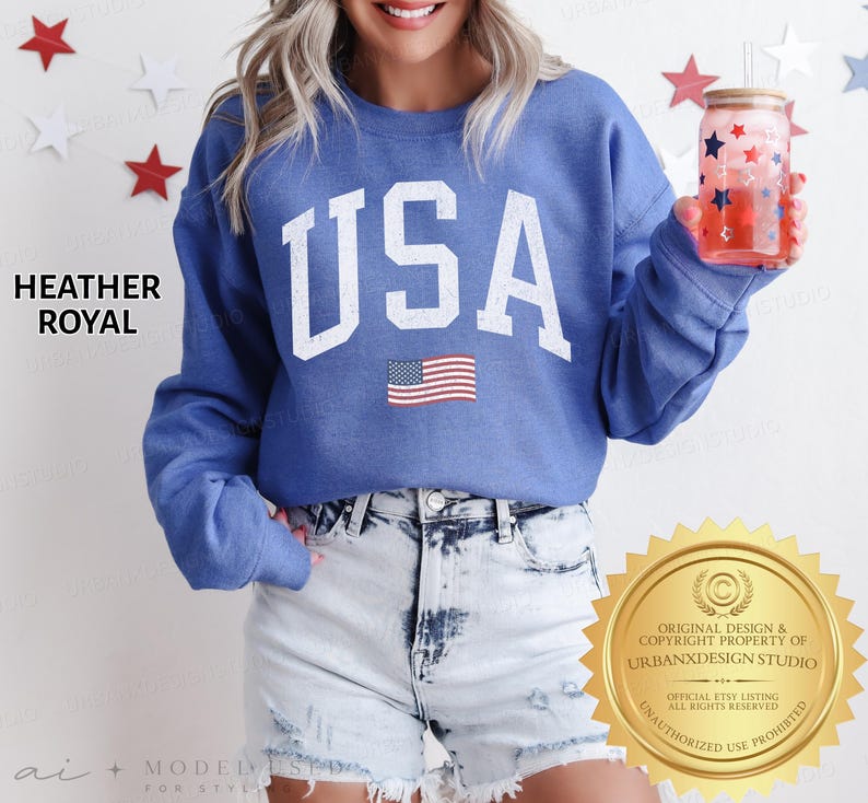 May include: A heather royal blue crewneck sweatshirt featuring the text USA in large white block letters with a distressed finish and a small American flag graphic below. The sweatshirt is paired with light blue acid-wash denim shorts.